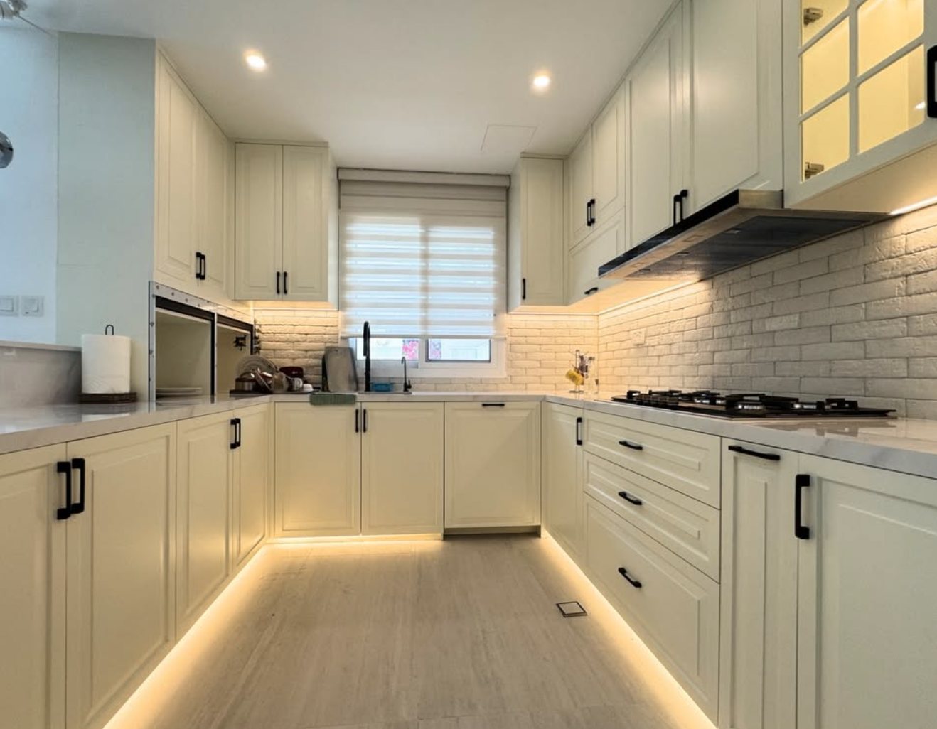 U shaped white kitchen with plinth lighting, brick backsplash and a roller blind